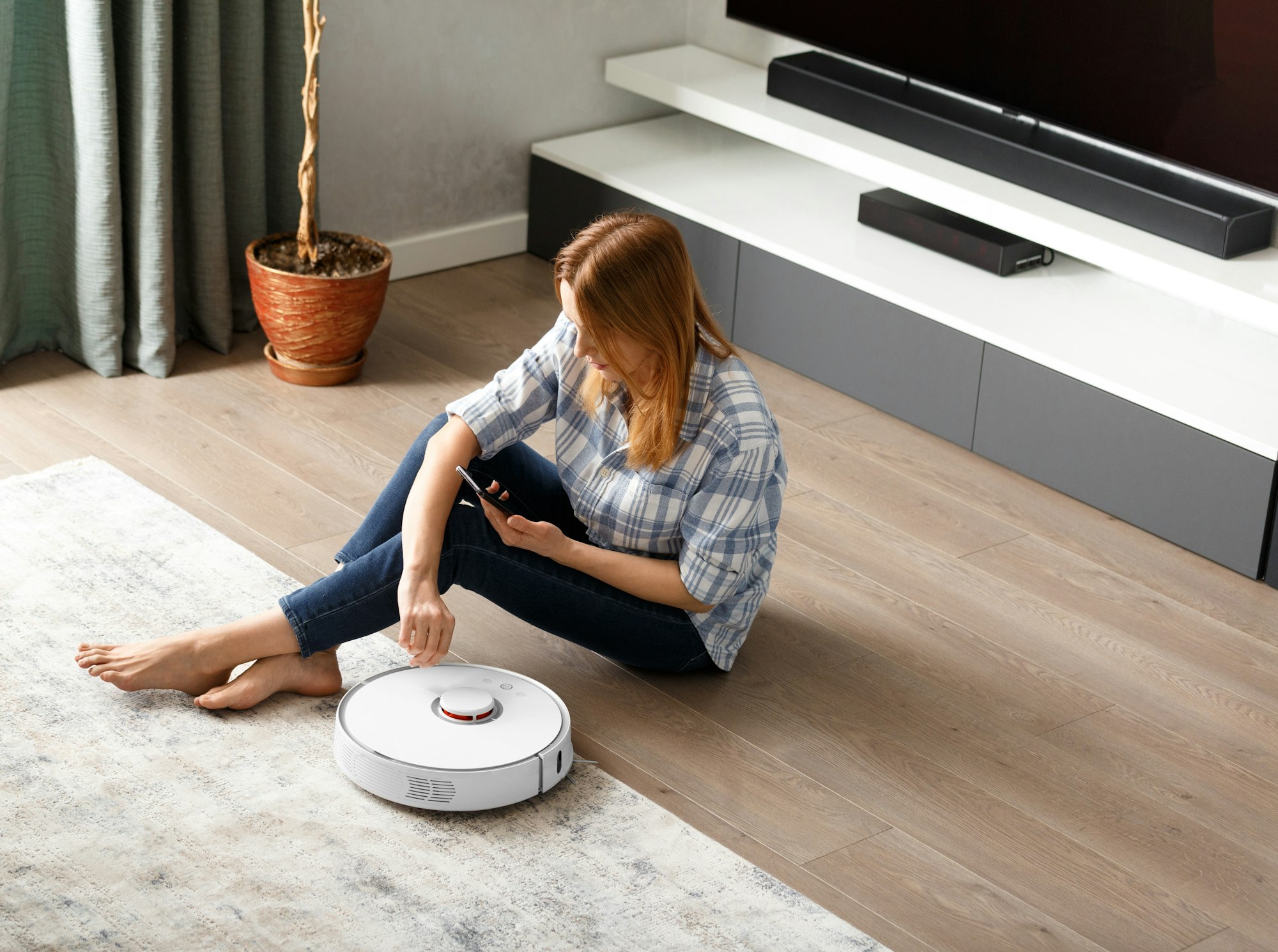 The girl sits on the floor and controls the robot vacuum cleaner using a smartphone, smart home.